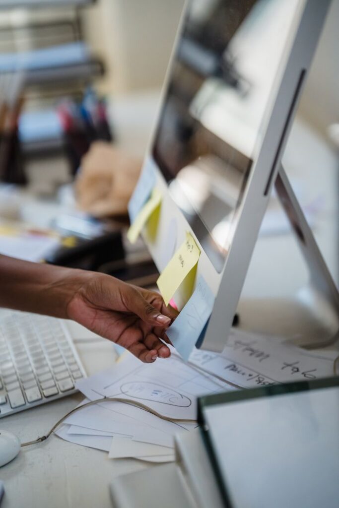 A focused shot of a hand placing a sticky note on a desktop monitor in an office setting.